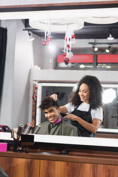 Sorridente uomo africano americano guardando specchio vicino parrucchiere con trimmer — Foto stock