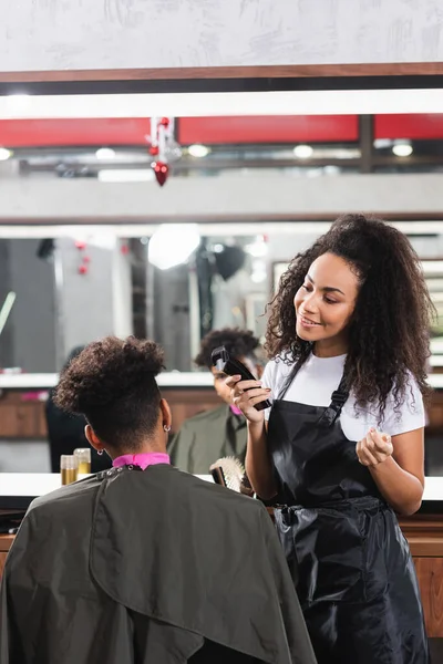 Усміхнений афроамериканський перукар тримає тример і дивиться на клієнта в салоні — стокове фото