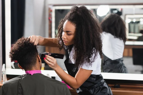 African american hairdresser in apron trimming head of client in salon — Stock Photo