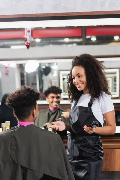 Smiling african american hairstylist with trimmer looking at client near mirror — Stock Photo