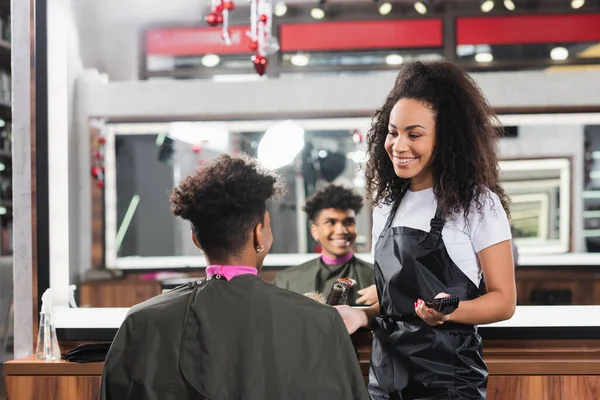 Cliente afroamericano sentado cerca de peluquero sonriente con trimmer en salón - foto de stock