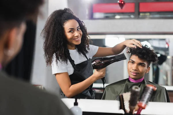 Cheerful african american hairstylist drying hair of young client on blurred foreground — Stock Photo