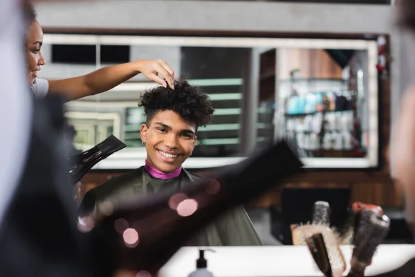 Smiling african american client sitting near hairdresser with hair dryer on blurred foreground — Stock Photo