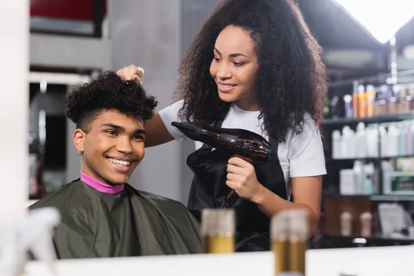 Cliente sorridente olhando espelho perto cabeleireiro com secador de cabelo no salão — Fotografia de Stock