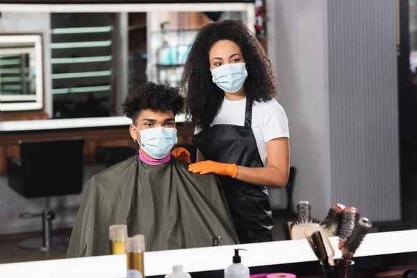 Parrucchiere afroamericano in maschera medica in piedi vicino al cliente in salone — Foto stock