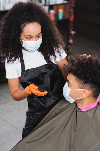 African american hairdresser in medical mask and latex gloves looking at blurred client — Stock Photo