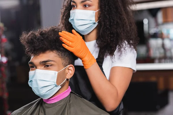 Client afro-américain en masque médical assis près du coiffeur sur fond flou — Photo de stock