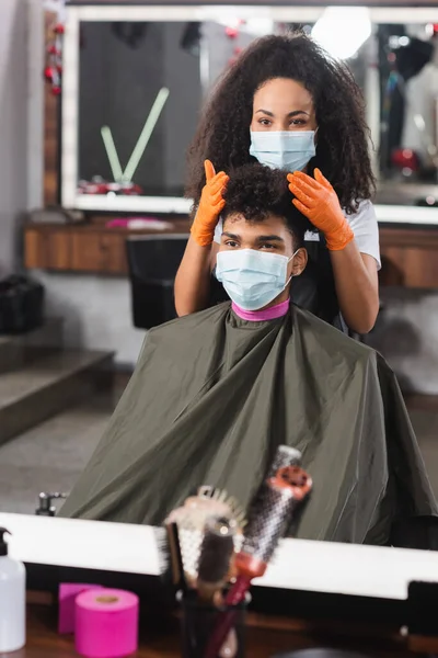 Coiffeur afro-américain en masque médical touchant les cheveux du client — Photo de stock