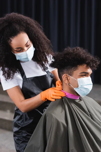 Coiffeur afro-américain en masque médical portant du papier collier sur le cou du client — Photo de stock