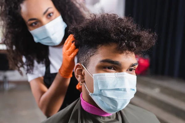 Hombre afroamericano en máscara médica sentado cerca de peluquero sobre fondo borroso - foto de stock