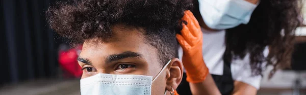 Vista recortada del hombre afroamericano sentado cerca de peluquero en máscara médica sobre fondo borroso, pancarta - foto de stock