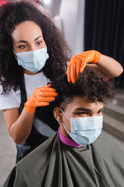 Африканский американский парикмахер в латексных перчатках на размытом фоне трогает волосы клиента в медицинской маске — стоковое фото