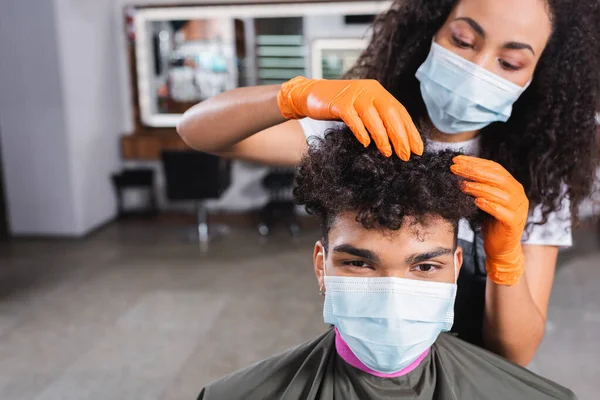 Cliente afroamericano en máscara médica sentado cerca de peluquero tocando el cabello sobre fondo borroso - foto de stock