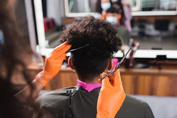 Ciseaux et peigne dans les mains du coiffeur afro-américain en gants de latex près du client — Photo de stock
