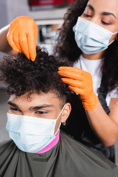 African american hairdresser in latex gloves on blurred background touching hair of client in medical mask — Stock Photo