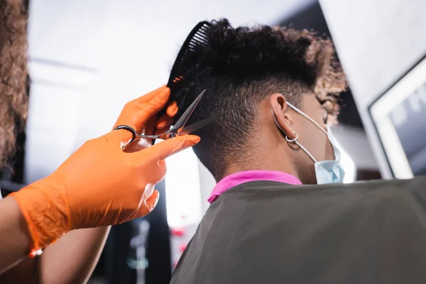 African american hairdresser in latex gloves holding scissors and comb near client during quarantine in salon — Stock Photo