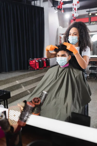 Peluquero afroamericano en guantes de látex y mascarilla médica cortando cabello de cliente en salón - foto de stock