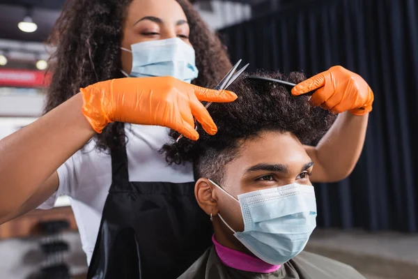 Африканский американский клиент в медицинской маске сидит рядом с парикмахером, стрижет волосы — стоковое фото