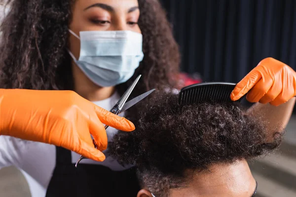 Scissors and comb in hands of african american hairstylist working with client in salon during quarantine — Stock Photo
