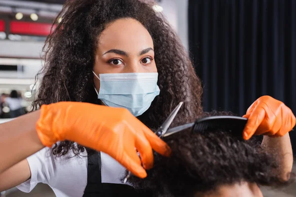 African american hairstylist in latex gloves and medical mask holding scissors and comb near hair of blurred man — Stock Photo