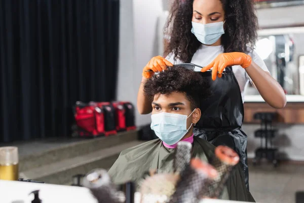 Peluquería afroamericana en guantes de látex y máscara médica cortando el cabello del cliente - foto de stock