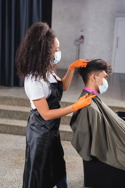 African american hairdresser in apron and protective mask combing hair of man in salon — Stock Photo