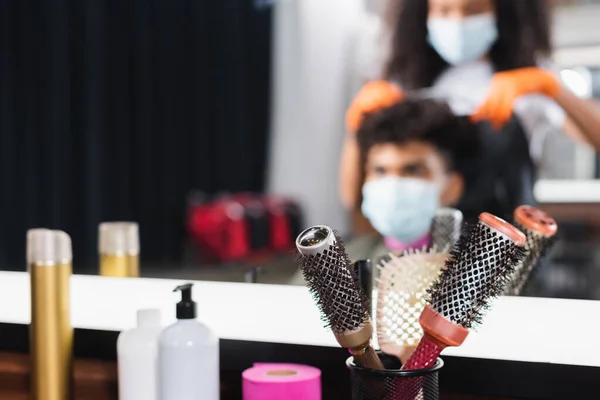 Cepillos cerca de espejo y peluquería afroamericana con cliente sobre fondo borroso en el salón - foto de stock