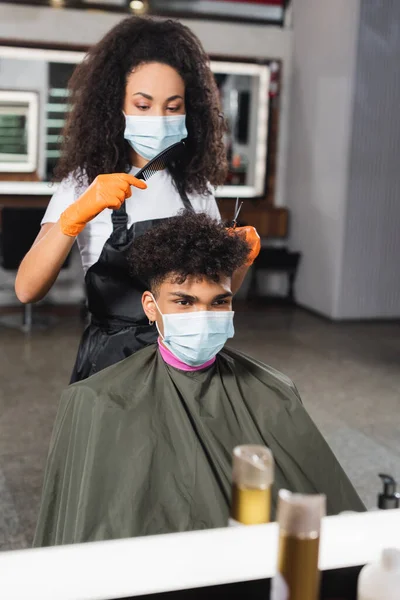 Coiffeur en masque médical tenant des ciseaux et peigne près du jeune homme afro-américain dans le salon — Photo de stock