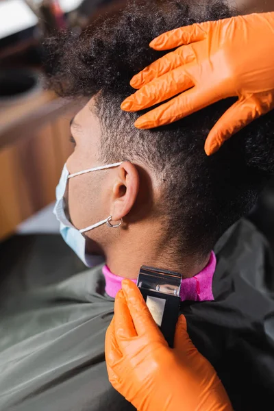 Peluquero en guantes de látex recortando cuello de hombre afroamericano en máscara médica y capa - foto de stock