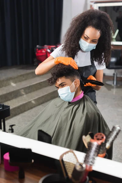 Afro-américain en masque médical assis près de coiffeur avec tondeuse — Photo de stock