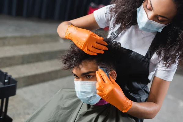 Peluquería afroamericana en guantes de látex y mascarilla médica recortando el cabello de un cliente joven - foto de stock