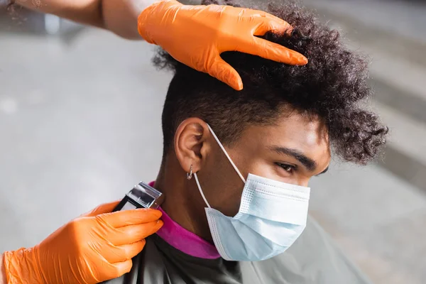 Cliente afroamericano in maschera medica seduto vicino parrucchiere in guanti di lattice utilizzando trimmer — Foto stock