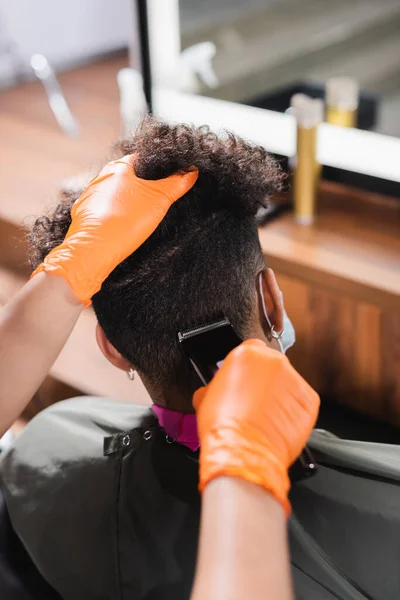 Peluquería en guantes de látex recortando la parte posterior de la cabeza del cliente afroamericano - foto de stock