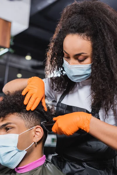 African american hairdresser in medical mask and gloves trimming hair of client on blurred foreground — Stock Photo