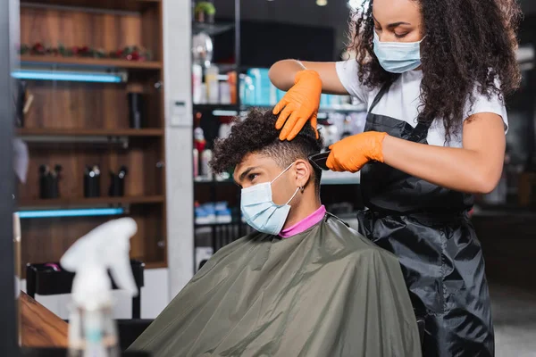 Cliente afroamericano en máscara médica situado cerca de peluquería con trimmer - foto de stock