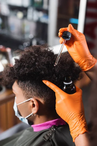 Парикмахер в латексных перчатках держит косметическое масло рядом с африканским американским клиентом в медицинской маске — стоковое фото