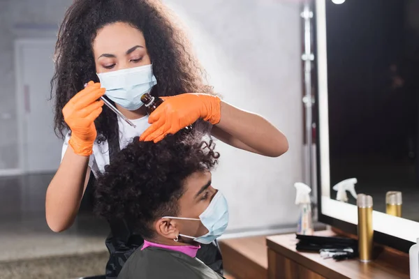 Coiffeur afro-américain en masque médical tenant de l'huile cosmétique près des cheveux du client dans le salon — Photo de stock