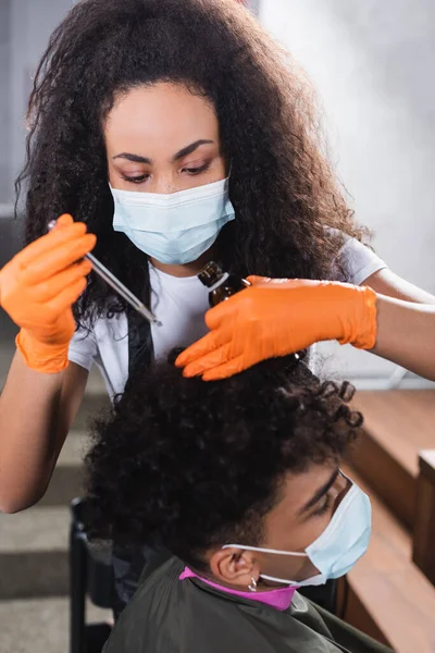 African american hairdresser in latex gloves holding cosmetic oil near hair of client on blurred foreground — Stock Photo