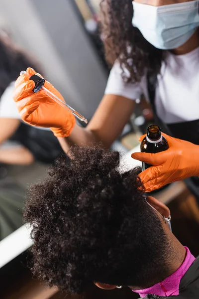 African american man near hairdresser on blurred foreground in latex gloves holding cosmetic oil — Stock Photo