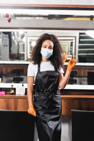 African american hairdresser in medical mask holding scissors and comb while pointing with finger in salon — Stock Photo