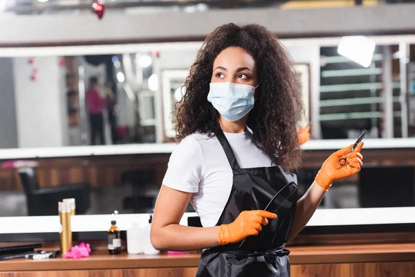 Jeune coiffeur afro-américain en tablier et masque médical tenant ciseaux et peigne — Photo de stock