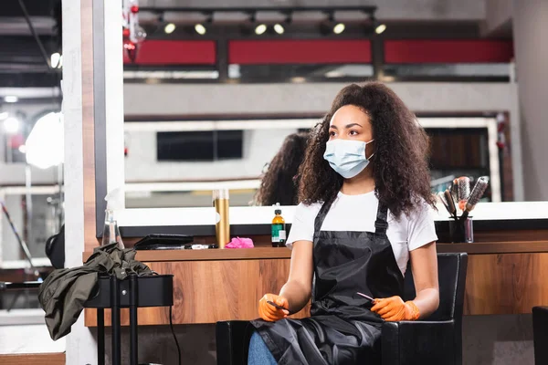 Peluquería afroamericana en máscara médica y guantes de látex con tijeras y peine - foto de stock