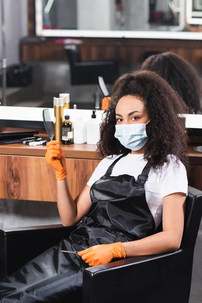 Afroamerikanischer Friseur in Schutzmaske hält Kamm und Schere im Stuhl — Stockfoto