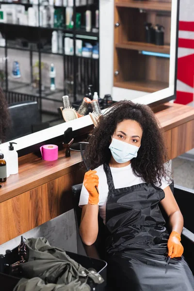 Африканський американський перукар в захисній масці і рукавичках, що тримають ножиці і гребінець біля дзеркала в салоні. — стокове фото