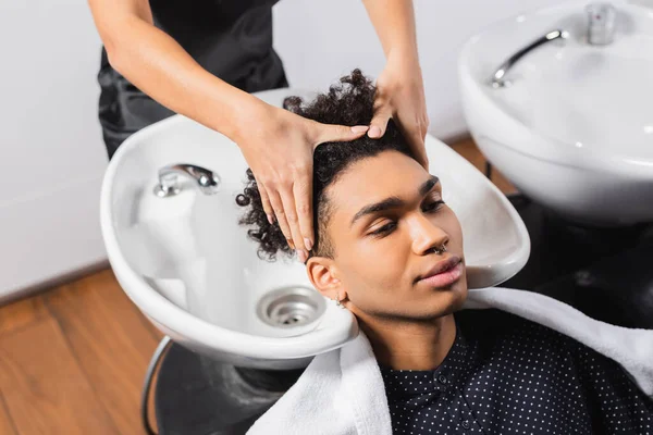 Africano americano homem sentado perto de cabeleireiro lavar o cabelo e pia no fundo borrado — Fotografia de Stock
