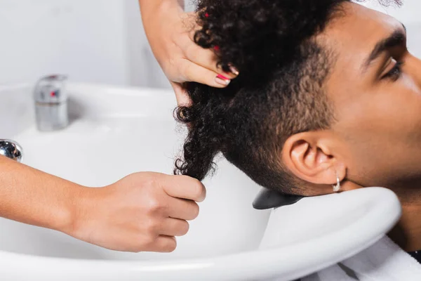 Vista recortada de peluquería lavando el cabello de un cliente afroamericano cerca del fregadero - foto de stock