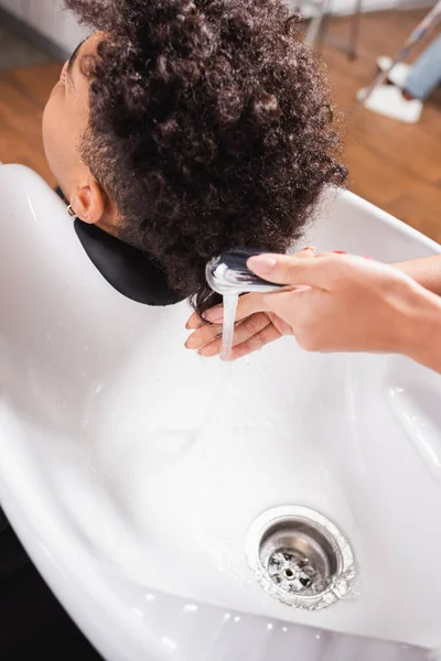 Hairdresser holding shower near african american client in salon — Stock Photo