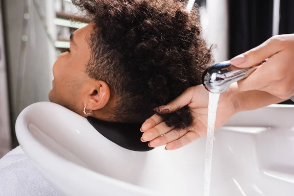 Hombre afroamericano sentado cerca de peluquería con ducha en el salón - foto de stock