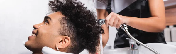 Client afro-américain souriant assis près du coiffeur avec douche sur fond flou, bannière — Photo de stock