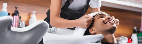 Friseur hält Handtuch in der Nähe des Kopfes eines lächelnden afrikanisch-amerikanischen Kunden und Waschbecken, Banner — Stockfoto